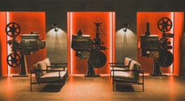 Drei historische Filmprojektoren vor rot beleuchteten Wänden und Sesseln